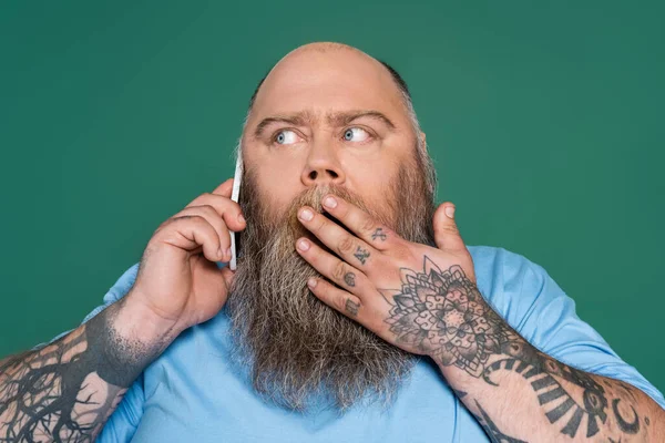 Sorprendido hombre tatuado que cubre la boca con la mano mientras habla en el teléfono inteligente aislado en verde - foto de stock