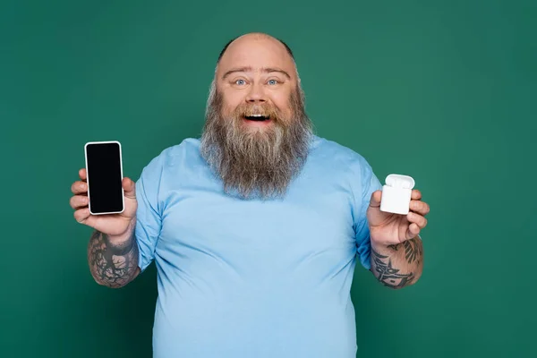 Hombre alegre más tamaño con teléfono móvil y auriculares inalámbricos aislados en verde - foto de stock