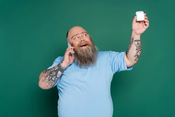 Cheerful plus size man talking on mobile phone and holding wireless earphones isolated on green — Stock Photo