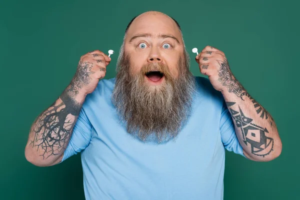 Amazed chubby man with beard holding wireless earphones isolated on green — Stock Photo