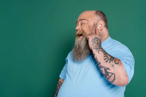 Pleased overweight man with closed eyes listening music in earphone and singing isolated on green — Stock Photo