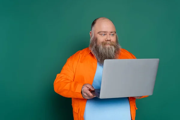 Barbuto uomo positivo con sovrappeso guardando computer portatile isolato su verde — Foto stock