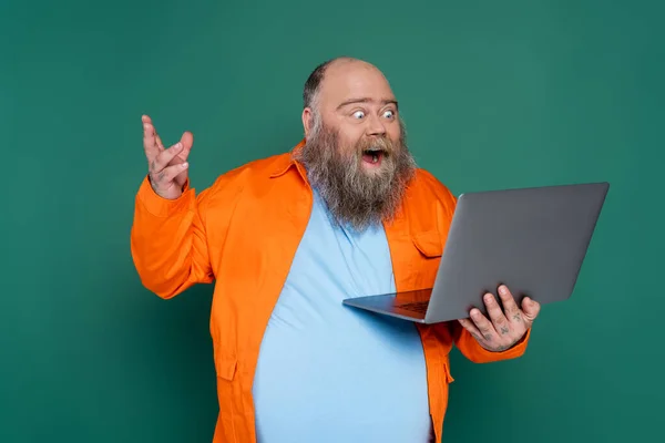 Erstaunt übergewichtiger Mann mit Bart schaut auf Laptop isoliert auf grün — Stockfoto