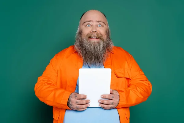 Asombrado y feliz hombre de tamaño grande con barba sosteniendo tableta digital aislado en verde - foto de stock