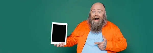 Взволнованный лишний вес человек указывая на цифровой планшет с чистым экраном изолированы на зеленый, баннер — стоковое фото