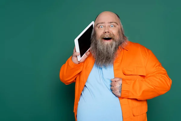 Bearded astonished man with overweight holding digital tablet near face isolated on green — Stockfoto