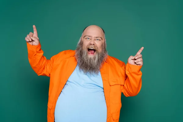 Thrilled overweight man screaming and pointing up with fingers isolated on green - foto de stock