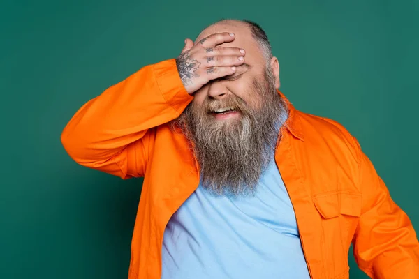 Barbudo hombre con sobrepeso riendo y cubriendo los ojos con la mano aislada en verde - foto de stock