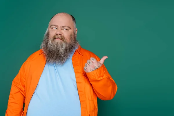 Bearded plus size man feeling disgusted and pointing with thumb isolated on green - foto de stock