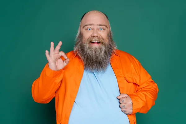 Excited plus size man in orange shirt showing okay sign isolated on green - foto de stock