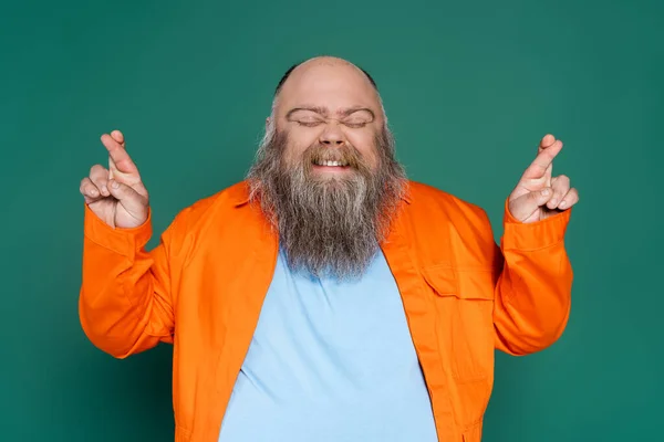 Hombre barbudo tenso con los ojos cerrados sosteniendo los dedos cruzados para la suerte aislado en verde - foto de stock