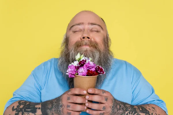 Pleased bearded man with overweight smelling aromatic flowers in paper cup isolated on yellow — Stock Photo