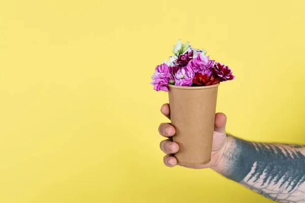 Частковий вид татуйованого чоловіка, що тримає одноразову чашку з натуральними квітами ізольовані на жовтому — стокове фото