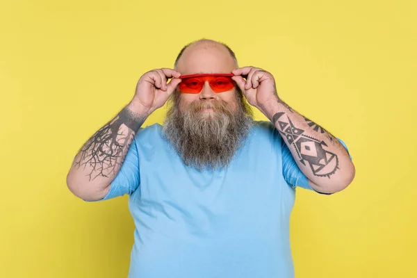 Gordito y hombre tatuado ajustando gafas de sol de moda mientras sonríe a la cámara aislada en amarillo - foto de stock