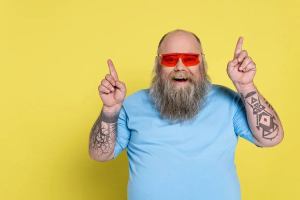 Bearded and cheerful overweight man in sunglasses pointing up with fingers isolated on yellow — Stockfoto