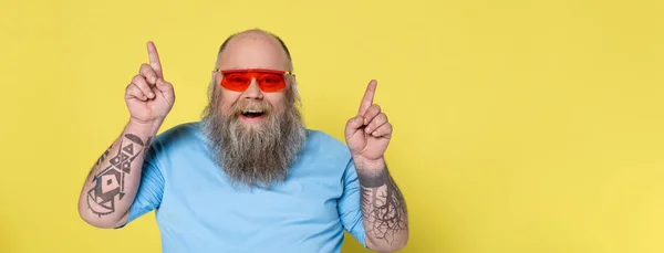 Joyful bearded man with overweight looking at camera and pointing up with fingers isolated on yellow, banner — Stock Photo