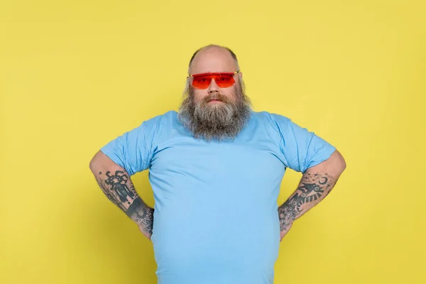 Homme barbu en surpoids dans des lunettes de soleil rouges debout akimbo isolé sur jaune — Photo de stock