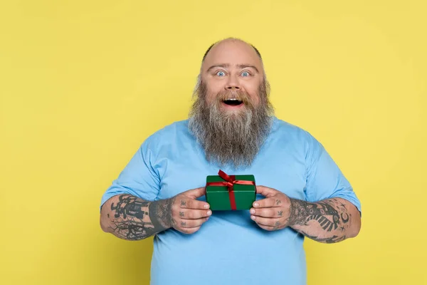 Homme barbu étonné avec excès de poids tenant boîte cadeau vert isolé sur jaune — Photo de stock
