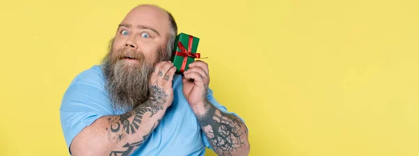 Surprised plus size man holding present while looking at camera isolated on yellow, banner — Stockfoto