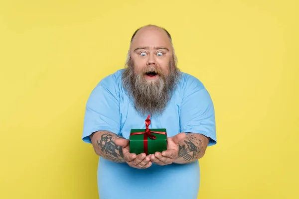 Homme en surpoids étonné avec barbe et tatouages regardant boîte cadeau vert isolé sur jaune — Photo de stock