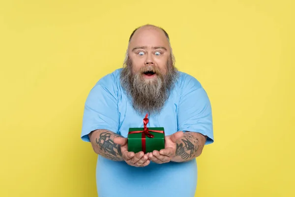 Astonished overweight man with beard and tattoos looking at green gift box isolated on yellow — Stock Photo