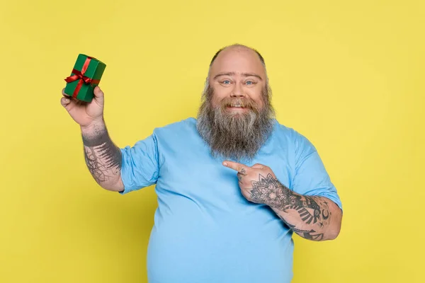 Uomo barbuto soddisfatto con sovrappeso puntando scatola regalo verde isolato su giallo — Foto stock