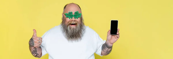 Excited plus size man in clover-shaped glasses holding smartphone and showing thumb up isolated on yellow, banner — Fotografia de Stock