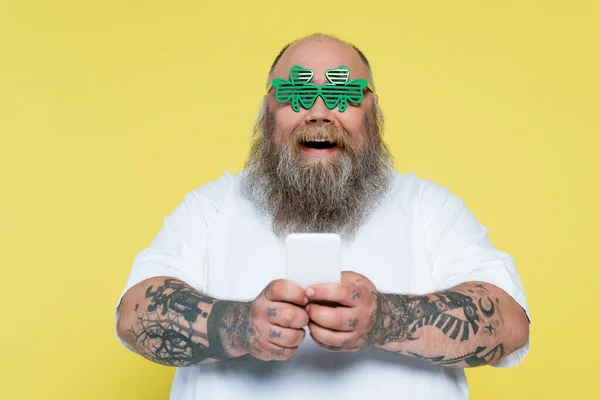 Amazed plus size man in clover-shaped glasses chatting on smartphone isolated on yellow - foto de stock