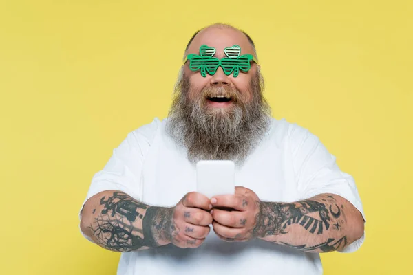 Amazed plus size man in clover-shaped glasses chatting on smartphone isolated on yellow — Stock Photo