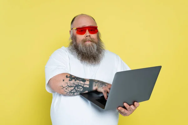 Elegante tatuado plus size homem em óculos de sol usando laptop isolado no amarelo — Fotografia de Stock