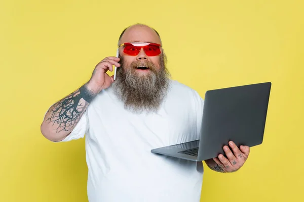 Excited overweight man in trendy sunglasses holding laptop and talking on smartphone isolated on yellow — Stock Photo