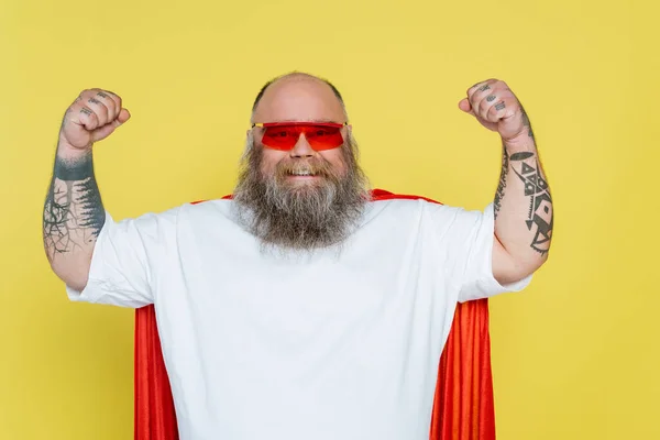 Homem forte e sobrepeso em óculos de sol vermelhos e capa de super-herói posando isolado no amarelo — Fotografia de Stock