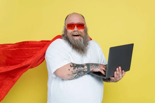 Thrilled overweight man in red sunglasses and superhero cloak using laptop isolated on yellow — Stock Photo