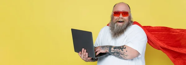 Cheerful overweight man in red sunglasses and superhero cloak using laptop isolated on yellow, banner — Stock Photo