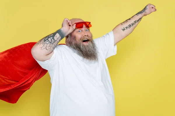 Hombre tatuado de talla grande en capa de superhéroe posando con la mano extendida aislada en amarillo - foto de stock