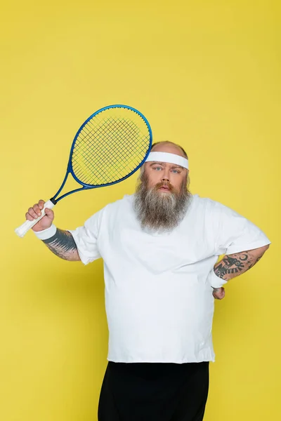 Barbudo jugador de tenis de tamaño grande de pie con la mano en la cintura y mirando a la cámara aislada en amarillo - foto de stock