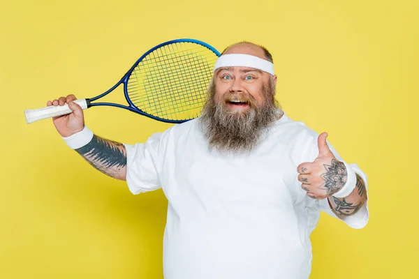 Gioioso uomo in sovrappeso con barba che tiene la racchetta da tennis e mostra il pollice in alto isolato sul giallo — Foto stock