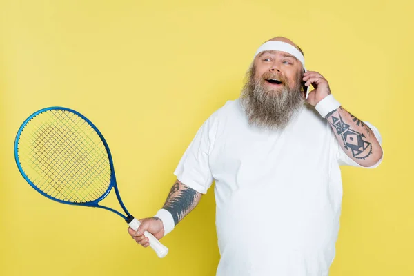Щасливий плюс розмір чоловік з тенісним ракеткою розмовляє на смартфоні ізольовано на жовтому — стокове фото
