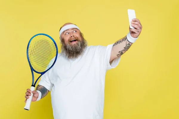 Hombre feliz y regordete tomando selfie con raqueta de tenis aislado en amarillo - foto de stock