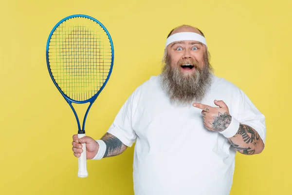 Amazed plus size man pointing at tennis racquet and looking at camera isolated on yellow — Stock Photo