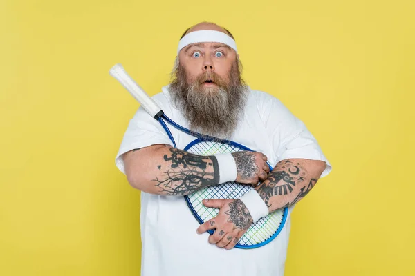 Shocked overweight man hugging tennis racquet and looking at camera isolated on yellow — Stockfoto