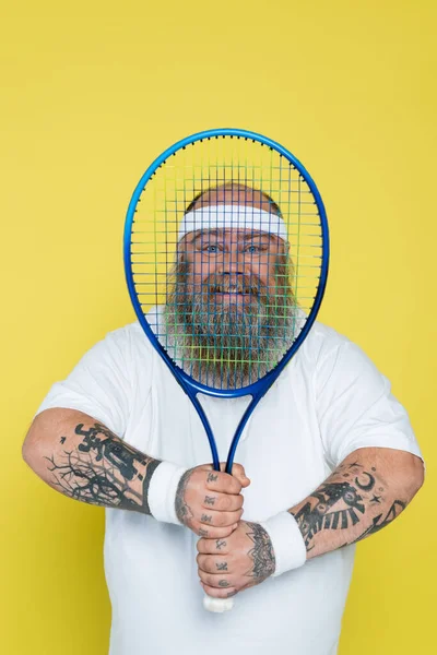 Heureux homme en surpoids avec barbe regardant caméra à travers raquette de tennis isolé sur jaune — Photo de stock