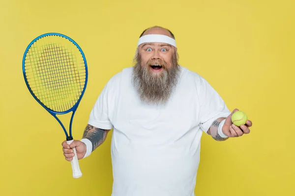 Asombrado hombre de tamaño grande con pelota de tenis y raqueta mirando a la cámara aislada en amarillo - foto de stock