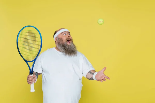 Surpreso homem com sobrepeso com raquete jogando com bola de tênis isolado no amarelo — Fotografia de Stock