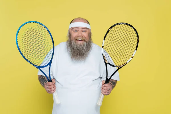 Pleased plus size man with beard holding tennis racquets isolated on yellow — Photo de stock