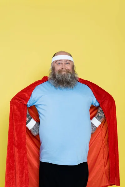 Plus size sportsman in superhero cloak and headband smiling at camera isolated on yellow — Photo de stock