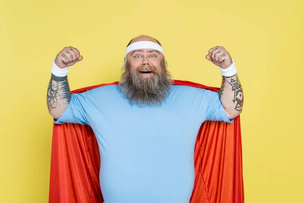 Bald and bearded overweight sportsman in superhero cloak showing power isolated on yellow — Foto stock