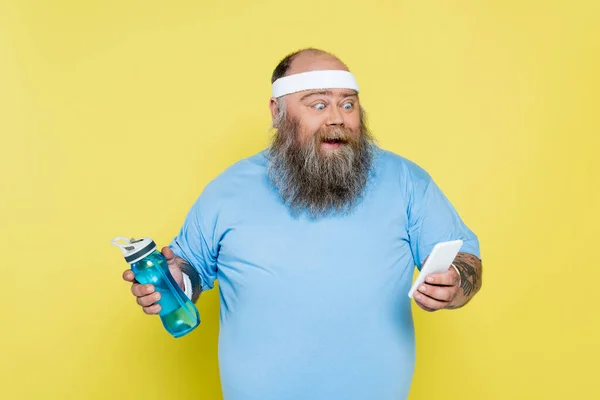 Homme en surpoids étonné avec bouteille de sport en utilisant un téléphone mobile isolé sur jaune — Photo de stock