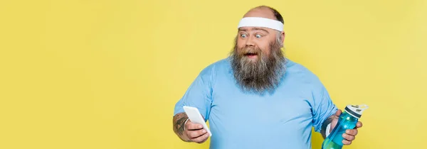 Amazed plus size man with sports bottle looking at smartphone isolated on yellow, banner — Foto stock