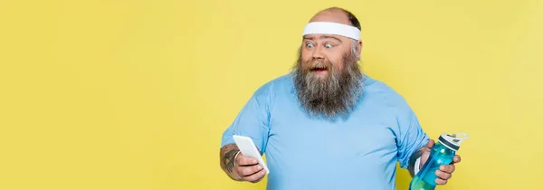 Изумленный плюс размер человека со спортивной бутылкой глядя на смартфон изолирован на желтый, баннер — Stock Photo